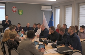 grupa siedzacych osob, na drugim planie stojacy mezczyzna z mikrofonem