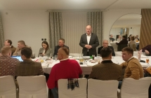 Grupa siedzacyh przy stole i stojacy mezczyzna