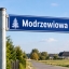 Znak z nazwa ulicy modrzewiowa. Bialy napis na niebieskim tle. w oddali wiejski krajobraz